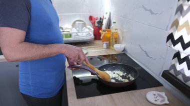 Adamın biri ocakta tavada yemek pişiriyor. Öğrenci soğan kızartıyor, yakın plan. Mavi bir gömlek giyiyor ve elinde ahşap bir kürek tutuyor. Mutfak çeşitli mutfak aletleri ve ev aletleriyle donatılmıştır.