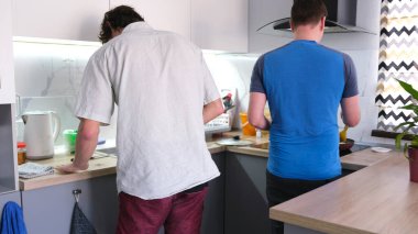 Lezzetli bir yemek pişiren eşcinsel bir çiftin arka planı. Mutfakta iki adam var, biri mavi bir gömlek giyiyor. Mutfakta çalışma masası ve saksı bitkisi var..