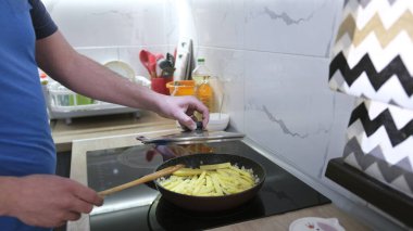 Patates kızartan bir adamın yakın çekimi. Adam ocakta yemek pişiriyor. Patates dilimlerini tahta bir kaşıkla karıştırıyor. Öğrenci kendi öğle yemeğini hazırlar. Tek bir adam.