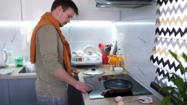 Beyaz bir adam yumurta kızartacak. Mutfak sobasının yanında duruyor. Mutfak iyi donatılmış. Adam turuncu bir kazak giyiyor ve işine odaklanmış durumda..