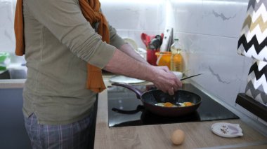 Ocakta tavada kızarmış yumurta pişiren beyaz bir adamın yakın çekimi. Mutfaktaki adam. Bekar hayatı. Mutfak çeşitli mutfak malzemeleriyle donatılmıştır.