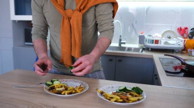 Mutfak masasında kız arkadaşı için lezzetli bir yemek hazırlayan bir adamın yakın çekimi. Salatalıkları dilimleyip kızarmış patatesle tabağa koyuyor. Mutfak çeşitli eşyalarla dolu.