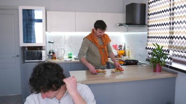 Çekici bir adam erkek arkadaşı için lezzetli ve sağlıklı bir akşam yemeği pişirdi. Erkek arkadaşı masada oturmuş lezzetli bir yemek bekliyor. Sıradan bir eşcinsel aile..