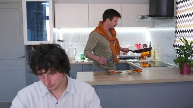 Bir adam mutfakta yemek pişirirken, diğeri masada oturuyor. Genç eşcinsel erkeklerin olduğu bir aile. Yurttaki iki oda arkadaşı için öğle yemeği. Yemek pişiriyorum.