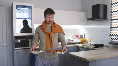 Turuncu kazaklı şık bir genç adam mutfak masasına tabak tabak taşıyor. İki kişilik yemek. Mutfak modern bir şekilde ışıklandırılmış ve dekore edilmiş..
