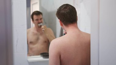 Diş bakımı. Adamın biri aynanın önünde dişlerini fırçalıyor. Ayna banyo duvarında asılıydı ve adam önünde duruyordu. Arkaya odaklan. Sabah tedavileri