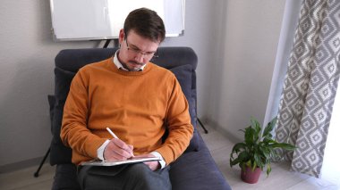 Gözlüklü ve turuncu kazaklı bir adam tablete bir şey yazıyor. Bu bir psikolog ya da öğretmen. Bir iş adamı, duvarında beyaz bir tahta ve yeşil bir saksı bitkisi olan bir odada mavi bir kanepede oturuyor.