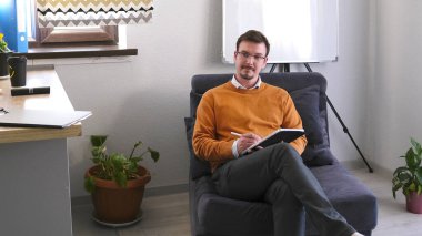 Psikolog 'un ofisi. Gözlüklü genç bir adam elinde bir defter ve kalemle mavi bir koltukta oturuyor. Turuncu bir kazak giyiyor ve yazmaya odaklanmış. Odanın tezgahı ve saksısı olan bir mutfak alanı var..