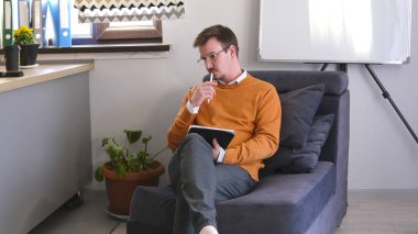 Düşünceli bir adam arkasında beyaz bir tahtayla mavi bir koltukta oturuyor. O bir tablet ve bir kalem tutuyor. Psikologun ofisi. Oda modern minimalist tarzda dekore edilmiş, saksı bitkisi ve masanın üzerinde vazo var..