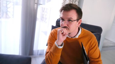 Gözlüklü ve turuncu kazaklı beyaz bir adam elinde tabletle mavi bir koltukta oturuyor. Psikolog düşüncelere daldı. Merkez ofisteki doktorun yakın çekimi.