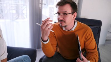 Turuncu kazaklı ciddi bir adam elinde beyaz bir kalemle tabletle koltukta oturuyor. Psikolog müşteriyle konuşur. Terapi. Düşünceli bir ruh hali içinde..