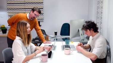 Pazarlama bölümünün başkanı ekibinin çalışmalarını denetliyor. Odada bir masada oturan iki kişi var ve onların arkasında turuncu kazaklı bir adam duruyor. Toplantı odası.