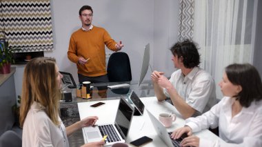 Ofiste buluşacağız. Bir adam konferans odasında bir grup insanın önünde sunum yapıyor. Grup, ikisi laptopları açık bir masada oturan dört kişiden oluşuyor..