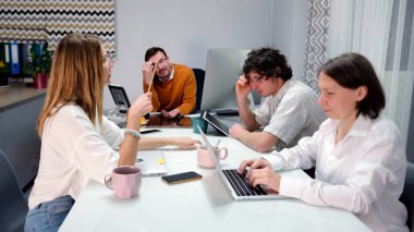 Ofis günleri. Bir grup genç insan bir masada oturuyor ve dizüstü bilgisayarları üzerinde çalışıyor. Beyin fırtınası. Atmosfer odaklanmış ve ciddi görünüyor.