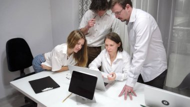 Bir grup genç şirket masasının başında toplandı ve bir dizüstü bilgisayara bakıyorlar. Sahne işbirlikçi ve odaklı. Öğrenciler ortak bir proje üzerinde çalışıyorlar.