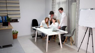 Küçük bir ev mini ofisi. Takım oluşturma ve işbirliği. Bir grup insan, yönetmenin beyaz masasında dizüstü bilgisayarlarıyla ekrana bakıyor. Sahne odaklı ve işbirlikçi.