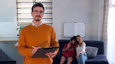 Başarılı bir psikolog portresi, mavi bir koltukta oturan genç evli bir çiftin arka planına karşı. Gözlüklü ve turuncu kazaklı bir adam elinde tabletle kanepenin önünde duruyor..