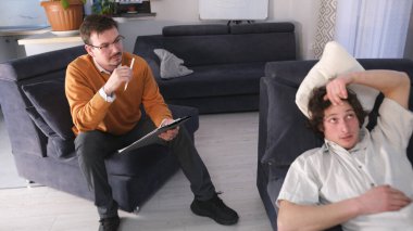 Genç bir adam psikoloğun seansına geldi. Psikoterapi. Turuncu kazaklı bir adam elinde tablet ve kalemle kanepede oturur ve hastasını dikkatle dinler. Uzman danışma