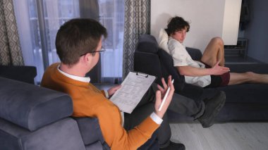 Profesyonel bir psikolog genç bir öğrenciyle psikoterapi seansı yapıyor. Çocukluk travmaları ve bağımlılıkların tedavisi. Turuncu kazaklı bir adam kanepede oturmuş beyaz gömlekli bir adamla konuşuyor.