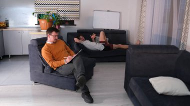 Psikanalist elinde bir tablet ve kalemle mavi bir koltukta oturuyor. Genç bir adam yakınlardaki başka bir kanepede yatar ve çocukluk travmalarından bahseder. Psikoterapi