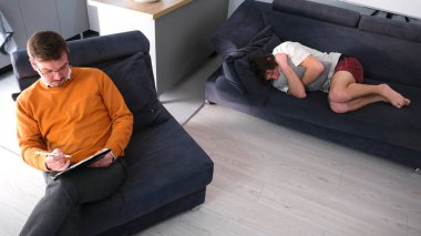 Yetenekli bir psikoloğun hizmetleri. Turuncu kazaklı ve gözlüklü bir psikanalist mavi koltukta otururken tablete bakıyor. Genç bir adam doktoruyla psikoterapi seansına geldi.