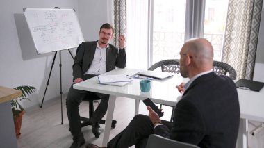 İki işadamı bir ofiste masa başında oturmuş bir şey tartışıyorlar. Bir tanesi elinde bir cep telefonu tutuyor. Sahne profesyonel ve odaklanmış. Noterin toplantı odası.