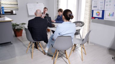 Meslektaşlar toplantı odasındaki brifinge gidiyorlar. Bir grup insan konferans odasında bir masada oturuyor. Takım elbiseli adamlar birbirleriyle konuşuyorlar ve masada birkaç dizüstü bilgisayar var..