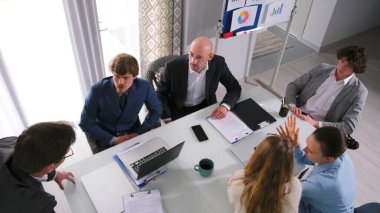 Konferans odasındaki bir masada oturan bir grup insanın üst görüntüsü. Hepsi iş kıyafetleri giyip şirketin geliştirme stratejisini tartışıyor. Sahne profesyonel ve odaklı.