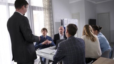 Şirketin erkek yöneticisi bir sunum yapıyor ve bir grup insan dikkatle dinliyor. Atmosfer ciddi ve odaklı. Bir grup insan bir masada oturuyor.