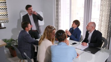 Ofiste verimliliği ve takım çalışmasını arttırma tartışması. Takım elbiseli ve gözlüklü bir koç beyaz tahtanın önünde danışmanlık yapıyor. Sahne profesyonel ve ciddi.