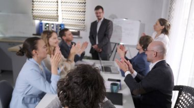 Ofis çalışanları başarılı bir rapor için meslektaşlarını tebrik ediyor. Bir grup insan ellerini çırpıyor ve beyaz tahtanın önünde duran mütevazı bir adama gülümsüyor. İş konsepti