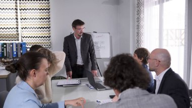 Şirketin müdürü bir konferans odasında bir grup insanın önünde duruyor. Adam takım elbise ve gözlük takıyor. Çalışanlar dizüstü bilgisayar ve kağıtlarla dolu bir masada oturuyorlar.