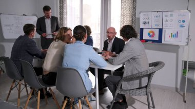 Bir grup yönetici konferans odasındaki masada oturarak iletişim kuruyor. Takım elbiseli bir adam sunum yapıyor. Sahne profesyonel ve odaklı.