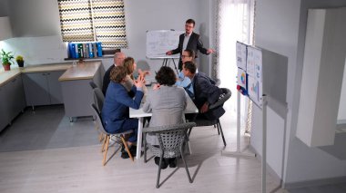 Şirketin danışmanı bir iş stratejisi geliştirmek için bir sunum yapıyor. Bir grup genç çalışan bir ofiste bir masada oturuyorlar. Sahne ciddi ve odaklanmış.