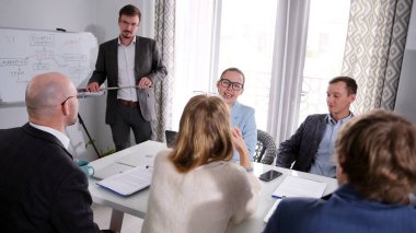 Şirket yönetim toplantısı konsepti. Bir grup iş arkadaşı bir sabah toplantısında bir masada oturuyor. Bir adam tahtanın önünde durup bir grupla konuşur..