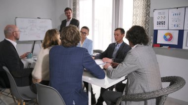 Bir grup insan konferans odasında bir masada oturuyor ve çalışanların işten çıkarılmasını tartışıyorlar. Ofis duvarında beyaz bir pano ve renkli grafiklerle dolu bir poster var..