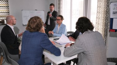 Akıl hocası genç bir işletme şirketinde ders veriyor. Bir grup insan bir toplantıda bir masada oturuyor. Bir adam ayakta bir grupla konuşuyor. Sahne profesyonel ve odaklı.