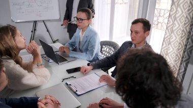 Bir grup genç iş adamı bir masada oturuyor ve bir proje üzerinde çalışıyorlar. Atmosfer ciddi ve odaklanmış durumda. Herkes dizüstü bilgisayarlarına ve evraklarına bakıyor.