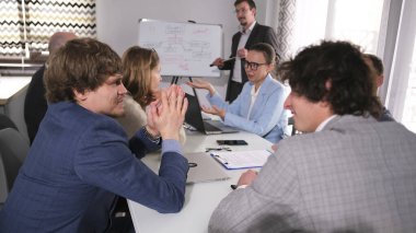 İş adamları yönetim kurulu toplantısında iletişim kurar. Bir grup insan bir masada oturuyor, bazıları takım elbiseli. Sahne ciddi ve odaklanmış.