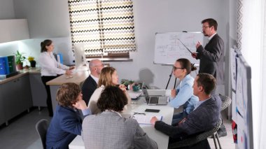 Bir grup iş adamı konferans odasında bir masada oturuyor. Koç beyaz tahtanın önünde duruyor ve bir sunum yapıyor. Sahne profesyonel ve odaklı.