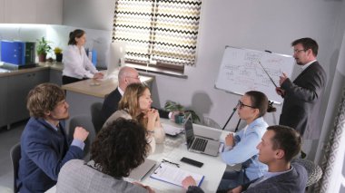 Bir misafir koç genç bir işletme şirketinde eğitim veriyor. Bir grup çalışan bir masada oturmuş yeni bir projenin çalışmalarını tartışıyorlar. Sahne profesyonel ve odaklı.