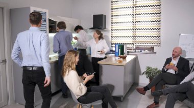 Bir grup iş arkadaşı iş yemeği için mutfakta toplandı. Aile ofisinde öğle yemeği. Olay yeri sıradan ve sosyal. Bazıları oturuyor, bazıları ayakta duruyor. Bir kadın sandalyede oturuyor ve telefonuna bakıyor..