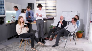 İş yemeği. Bir grup şirket yöneticisi mutfakta toplandı, bazıları sandviç ve salata yerken, diğerleri kendilerine kahve koyuyorlar. Atmosfer sakin görünüyor.