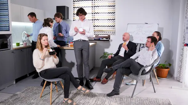 İş yemeği. Bir grup şirket yöneticisi mutfakta toplandı, bazıları sandviç ve salata yerken, diğerleri kendilerine kahve koyuyorlar. Atmosfer sakin görünüyor.