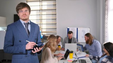 Takım çalışması. Takım elbiseli ve gözlüklü ciddi bir yönetici konferans odasında bir grup insanın önünde duruyor. Elinde bir kalem ve tablet var..