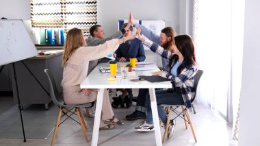 Takım oluşturma ve işbirliği. Bir grup yaratıcı insan toplantı odasında bir masada oturup birbirlerine beşlik çakıyorlar. Sahne olumlu ve dostane.