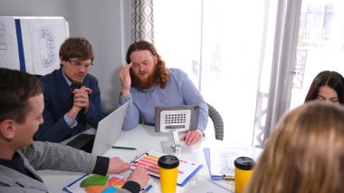 Yakışıklı kızıl saçlı bir adam patronun özel asistanıdır, ona önemli bilgiler verir. Bir grup insan laptopları ve kahve bardaklarıyla bir masada oturuyor..