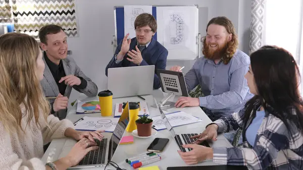 Takımda dostane ve olumlu bir atmosfer var. Bir grup çalışan bir masada oturuyor ve birlikte çalışıyorlar. Masa dizüstü bilgisayarlar, kahve fincanları, kağıtlar vs. ile dolu..