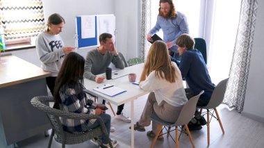 Kreatif direktör sabah brifingi için ekibini topladı. Bir grup insan bir aile ofisinin konferans odasında bir masada oturuyor..