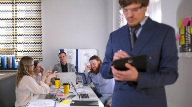 Takım elbiseli bir adam konferans odasında bir grup insanın önünde duruyor. Elinde bir tablet tutuyor ve bir şeyler yazıyor. Bir grup insan dizüstü bilgisayarlarla bir masada oturuyor.